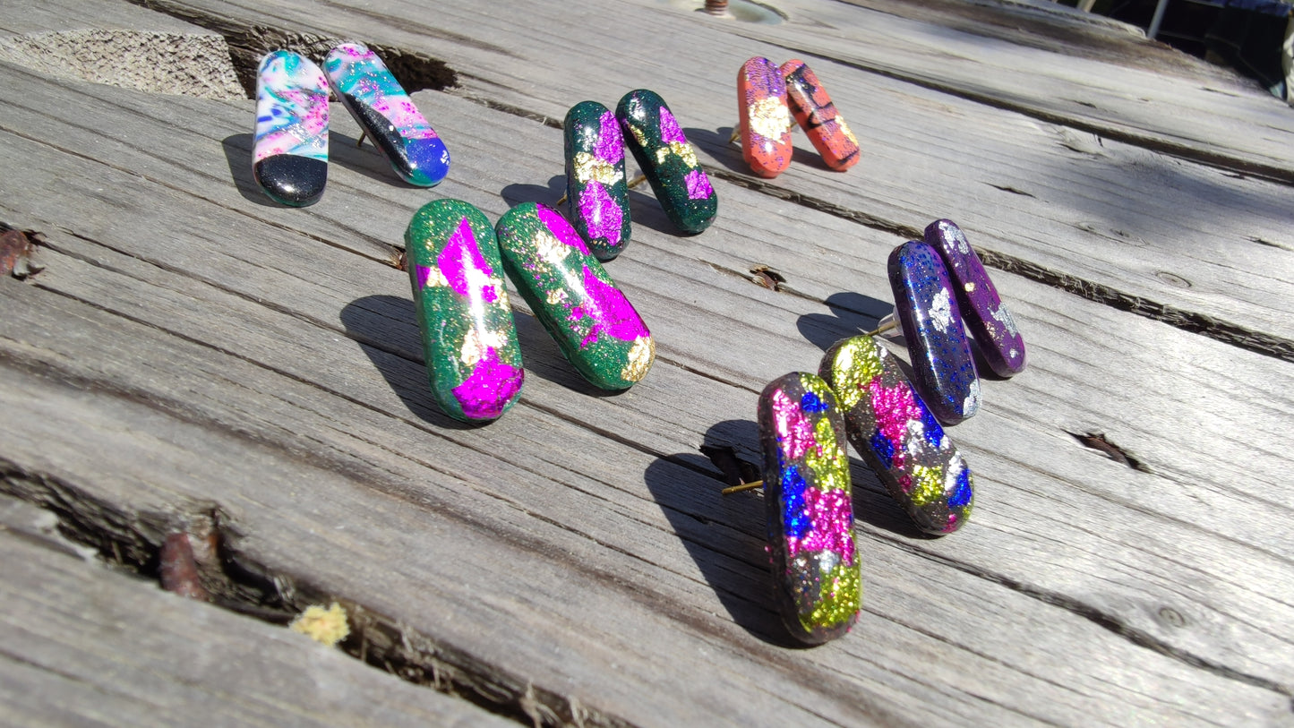 Boucles d'oreilles capsule Astral marbrées vert sapin éclats fuchsia et or, argile polymère résine UV, posées sur bois au soleil
